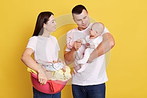 Hipster father and mother posing with thir child isolated over yellow background, attractive woman holding basin with clothing,