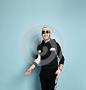 Hipster fellow in cap, black tracksuit and sunglasses. He laughing and pointing at something, posing on blue background. Close up