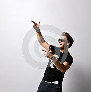Hipster fellow in sunglasses, black t-shirt with print, bracelets. He is pointing at someone, posing isolated on white. Close up