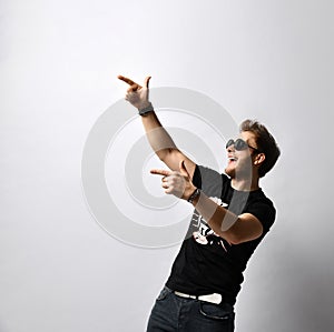 Hipster fellow in sunglasses, black t-shirt with print, bracelets. He is pointing at someone, posing isolated on white. Close up