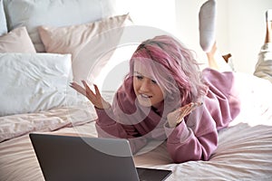 Hipster gen z teen girl with pink hair video calling on laptop lying in bed.