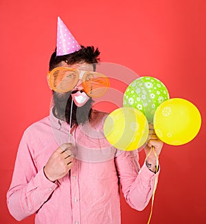 Hipster in giant sunglasses celebrating. Man with beard on cheerful face holds smiling mouth on stick, red background