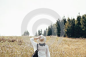 Hipster girl with backpack traveling in sunny mountains, walking in sunny wildflower meadow. Stylish woman in hat exploring and