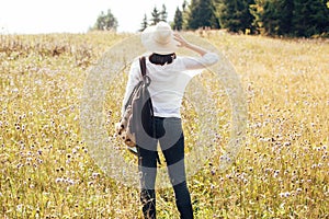 Hipster girl with backpack traveling in sunny mountains, walking in sunny wildflower meadow. Stylish woman in hat exploring and