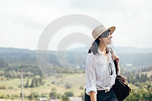 Hipster girl with backpack traveling on top of sunny mountain, walking on hills with woods. Stylish woman smiling enjoying hiking