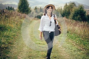 Hipster girl with backpack traveling on top of sunny mountain, walking on hills with woods. Stylish woman smiling enjoying hiking