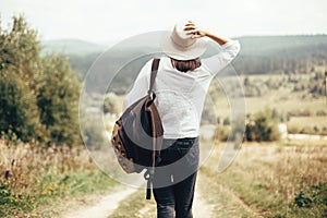 Hipster girl with backpack traveling and walking on top of sunny mountain and looking at hills and woods. Stylish woman in hat