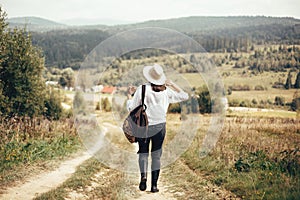 Hipster girl with backpack traveling and walking on top of sunny mountain and looking at hills and woods. Stylish woman in hat