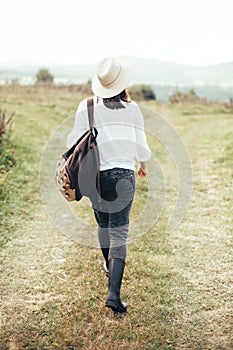 Hipster girl with backpack traveling and walking on top of sunny mountain and looking at hills and woods. Stylish woman in hat