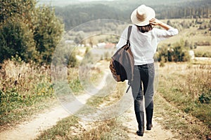 Hipster girl with backpack traveling and walking on top of sunny mountain and looking at sky and woods. Stylish woman in hat