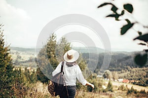 Hipster girl with backpack traveling and walking on top of sunny mountain and looking at sky and woods. Stylish woman in hat