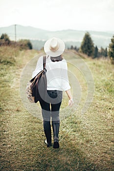 Hipster girl with backpack traveling and walking on top of sunny mountain and looking at sky and woods. Stylish woman in hat