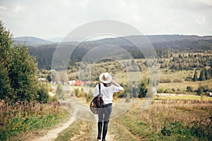 Hipster girl with backpack traveling and walking on top of sunny mountain and looking at sky and woods. Stylish woman in hat