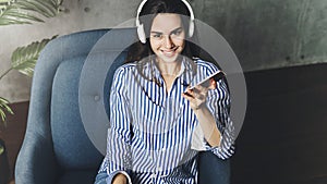 Hipster girl in earphones listening to music in online app sitting on sofa in her own apartment. Young lady downloading audio apps