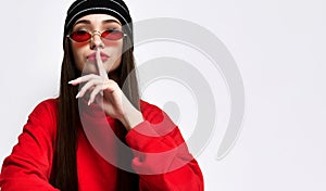 Hipster girl in a fashionable black hat headdress with long hair, showing hush, shhh, secrecy, silent sign, on light studio wall