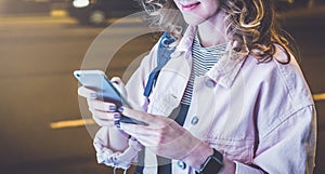 Hipster girl in glasses stands on night street and uses smartphone, touching screen with finger.