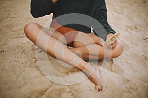 Hipster girl holding herb and sitting on beach, cropped view on tanned legs. Stylish boho woman in modern swimsuit and sweater