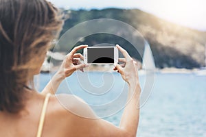 Hipster girl make photo on smart phone in coastline, mock up blank screen. Traveler using in female hand mobile on background