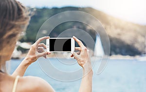 Hipster girl make photo on smart phone in coastline, mock up blank screen. Traveler using in female hand mobile on background