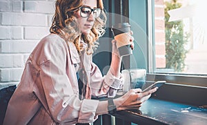 Hipster girl sitting in cafe at black table, drinking coffee and using smartphone. Businesswoman working online.