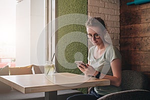 Hipster girl is sitting in cafe at table near window and looking on screen of smartphone.Young businesswoman working