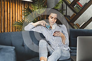 Hipster girl sitting in sofa and texting on smartphone. Entertaining and educating content online. Female communicating in social