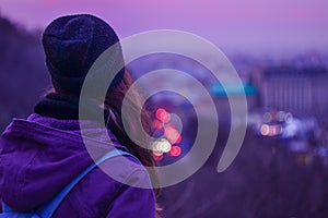 Hipster girl traveler looking at winter evening cityscape, purple sky and blurred city lights