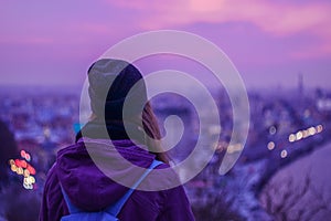 Hipster girl traveler looking at winter evening cityscape, purple violet sky and city lights