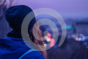 Hipster girl traveler looking at winter evening cityscape, violet sky and blurred city lights