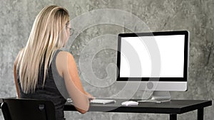 Hipster girl in trendy glasses sits at table in front of computer working. White Display.