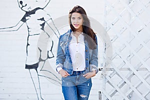 Hipster girl wearing blank white shirt, jeans and jeans jacket posing against street wall. Minimalist urban clothing