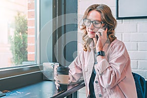 Hipster girl wearing glasses and pink denim jacket sitting in coffee shop near window