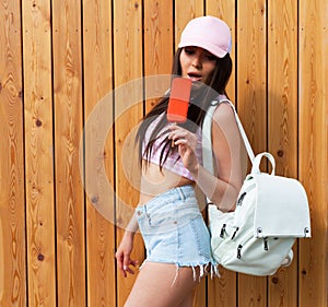 Hipster girl wearing pink top, jeans and baseball cap with white backpack posing against wood street wall, Enjoys ice cream. Urban