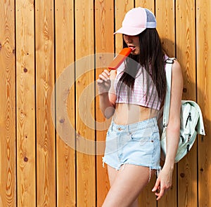 Hipster girl wearing pink top, jeans and baseball cap with white backpack posing against wood street wall, Enjoys ice cream. Urban