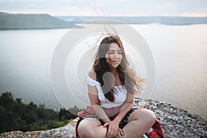 Hipster girl with windy hair sitting and meditating on top of rock mountain with beautiful view on river. Atmospheric calm moment