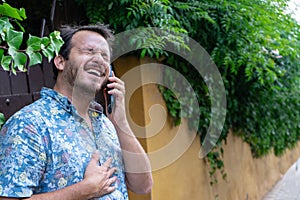 Hipster guy bearded laughing in the street while speak in a phone call. Young adult talking in cellular with a friend, listening a