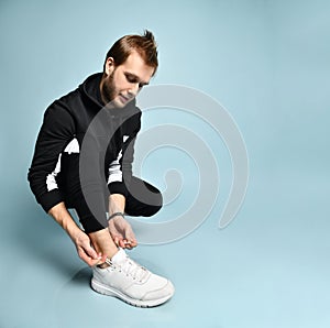 Hipster guy in black tracksuit, bracelet and sneakers. He is squatting and tying shoelaces, posing on blue background. Close up