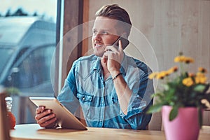 Hipster guy with a stylish haircut and beard sits at a table in a roadside cafe, talking on the phone and holds a tablet