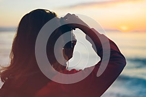 Hipster hiker tourist with backpack taking photo of amazing seascape sunset on camera on background blue sea, photographer enjoyin