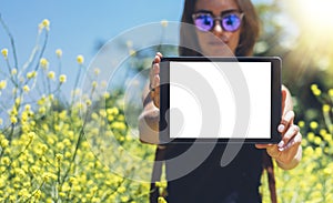 Hipster holding in hands tablet computer. Girl traveler with sunglasses using gadget on sun flare and yellow flowers background
