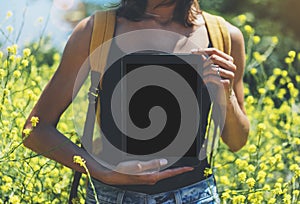 Hipster holding in hands tablet computer, mockup of blank empty screen. Girl traveler using gadget on sun flare and yellow flowers