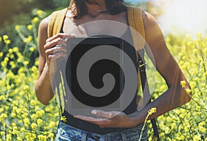 Hipster holding in hands tablet computer, mockup of blank empty screen. Girl traveler using gadget on sun flare and yellow flowers