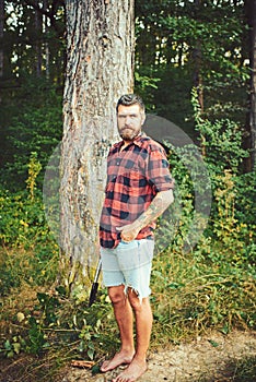 Hipster with long beard on natural green landscape. Bearded man with shovel in forest. Tourist in plaid shirt and jean