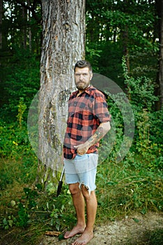 Hipster with long beard on natural green landscape. Bearded man with shovel in forest. Tourist in plaid shirt and jean