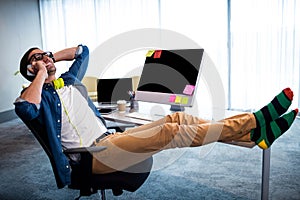 Hipster man calling with smartphone while relaxing at computer desk