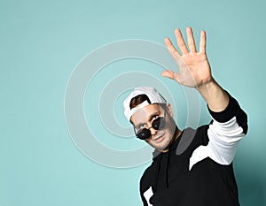 Hipster man in cap, black tracksuit, bracelet and sunglasses. He raised his palm up saying hi, posing on blue background. Close up