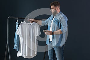 Hipster man dressed in denim shirt and jeans, stands indoors and looks at white T-shirt on clothes hanger in his hands.