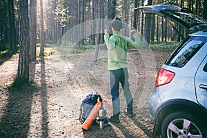 Hipster man in felt hat has just took backpack and Ñamping gear from car trunk after road trip, stretches and admires pine forest