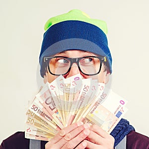 Hipster man in glasses covered face with money, looking aside. Guy in hat having fan of money in hand on white wall. Man saving