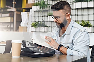 Hipster man in glasses is sitting in cafe, reading notes in notebook. On table is laptop,cup of coffee, instant camera.